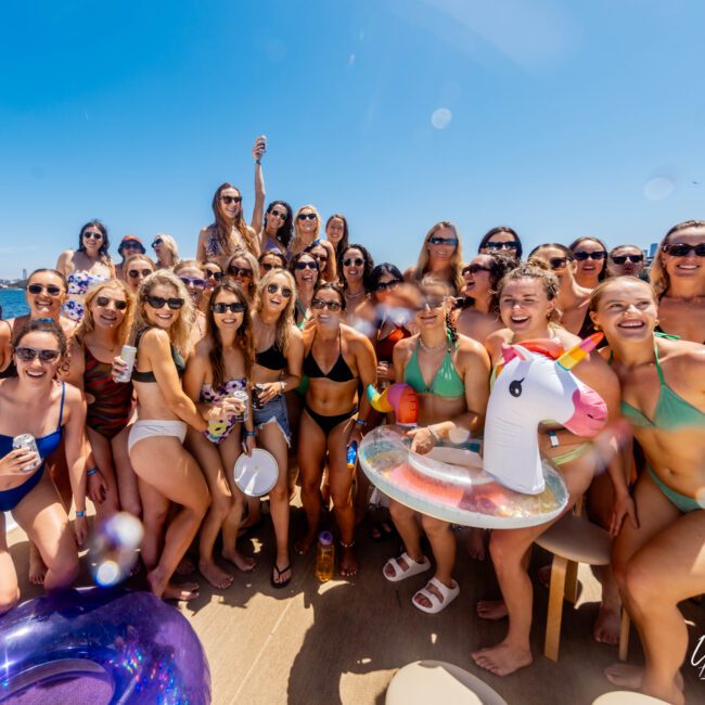 A large group of women in swimsuits gather for a photo on a boat under clear blue skies. Many are holding drinks, and one woman holds an inflatable unicorn. They all appear to be enjoying a sunny day on the water together. A logo in the corner reads "Luxury Yacht Rentals Sydney.