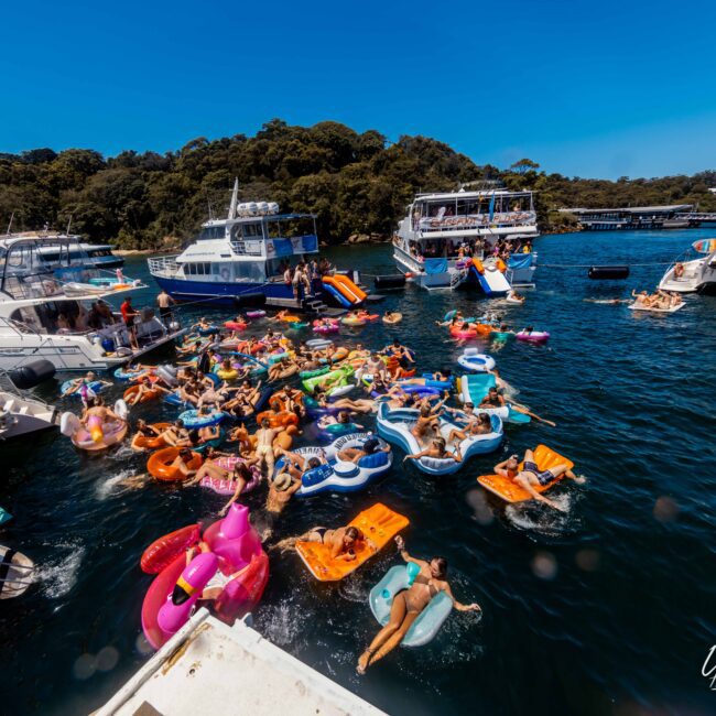 A lively scene at a yacht party in Sydney Harbour with multiple people floating on colorful inflatables in the water. Several luxury yachts are anchored nearby, with wooded hills in the background under a clear blue sky. A logo reading "The Yacht Social Club" is seen in the bottom right corner.