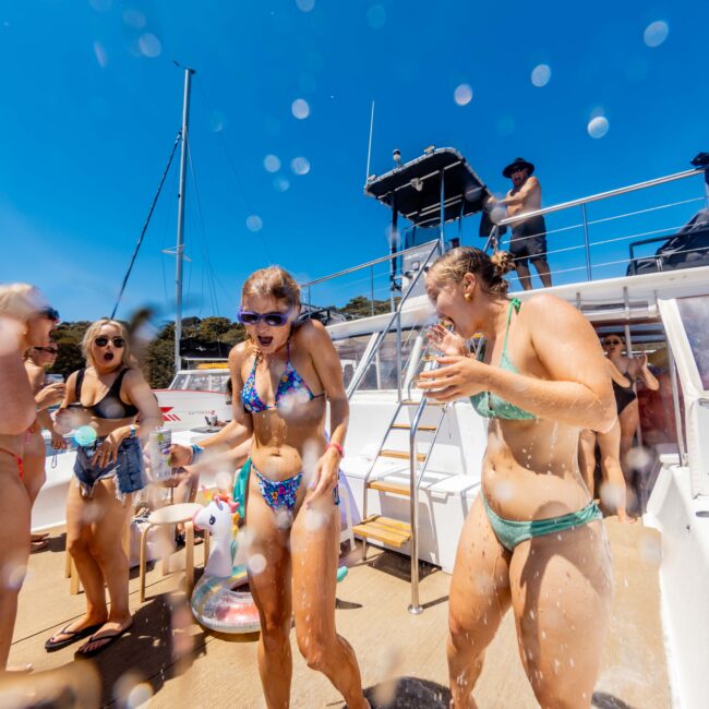 A group of people in swimsuits enjoy a sunny day on a yacht. Water splashes around them as they laugh and smile. The background shows a clear blue sky and part of the boat's upper deck, where more people are visible. "Boat Parties Sydney The Yacht Social Club" is written in the bottom right corner.