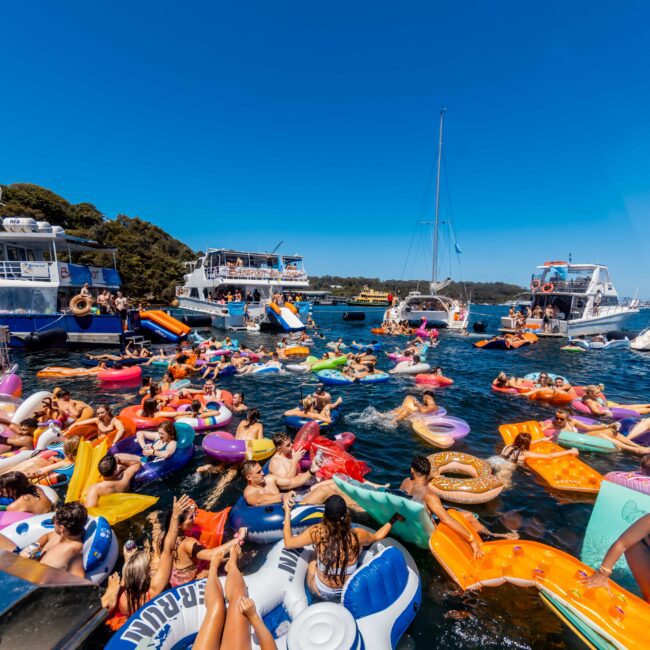 A lively scene of people enjoying a sunny day on the lake, floating on colorful inflatables. There are various pool floats like unicorns, pizza slices, and flamingos. Several boats are anchored nearby with more people onboard, reminiscent of The Yacht Social Club Sydney Boat Hire experience amidst lush green trees in the background.