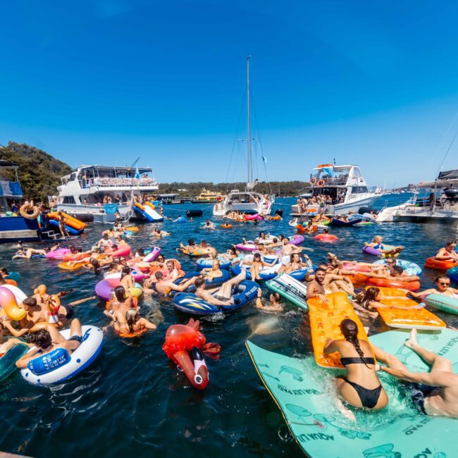 A lively scene of people on colorful inflatable floats enjoying a sunny day in the water. Several boats from The Yacht Social Club Sydney Boat Hire are anchored nearby, and the blue sky contrasts with the vibrant hues of the inflatables. The atmosphere is festive, with people relaxing and socializing in the sea.