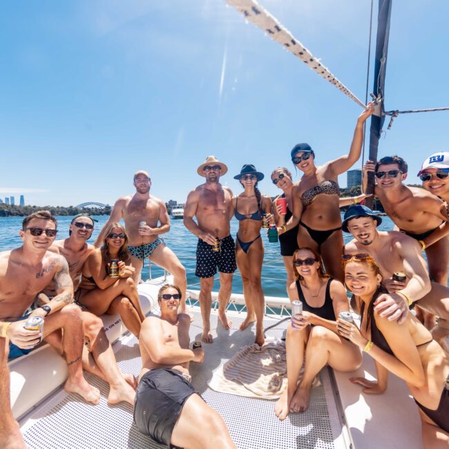 A group of people are enjoying a sunny day on a yacht during The Yacht Social Club Event. They are casually dressed in swimwear, sunglasses, and hats, and are smiling at the camera. The scene is bright with clear blue water and a city skyline in the background.