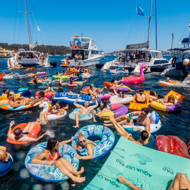 A lively gathering of people enjoying a sunny day on a lake. They are floating on colorful inflatables, including a giant pink flamingo and various other shapes, surrounded by boats. Blue skies and clear waters set the scene for a fun summer event hosted by The Yacht Social Club Sydney Boat Hire.