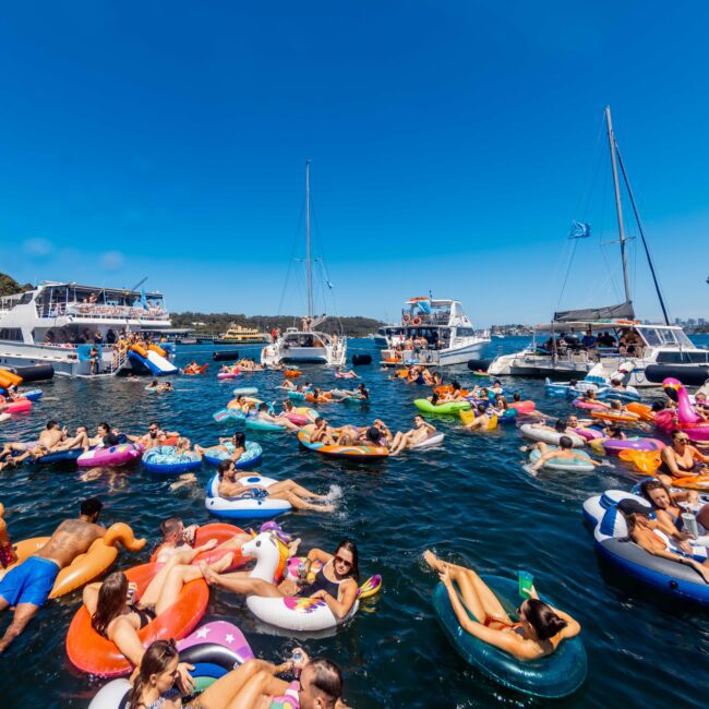 A large group of people are enjoying a sunny day on the lake, floating on colorful inflatables. Several boats and yachts from Sydney Harbour Boat Hire The Yacht Social Club are anchored nearby, creating a lively and festive atmosphere. The clear sky and trees in the background add to the vibrant scene.