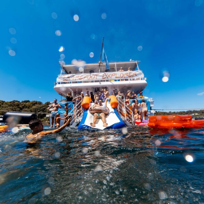 People enjoying a sunny day on the water, some aboard a large boat labeled "Seafood Explorer," and others nearby. One person is sliding into the water from a waterslide attached to the boat, surrounded by various flotation devices. Experience Boat Parties Sydney The Yacht Social Club for unforgettable fun.