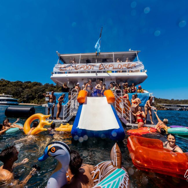 A lively party scene on a boat named "Barefoot Explorer," hosted by The Yacht Social Club Sydney Boat Hire, features people enjoying a large blue and white slide that extends into the water. Many revelers are in swimsuits, floating on colorful inflatables, with a backdrop of trees and other boats in the water.