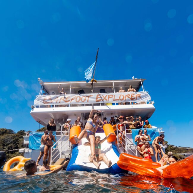 A group of people on a double-deck yacht enjoy a sunny day. Some are swimming with large inflatable toys, while others socialize on the "Barefoot Explorer." A person slides off the water slide into the sea. The logo of Sydney Harbour Boat Hire The Yacht Social Club is visible.