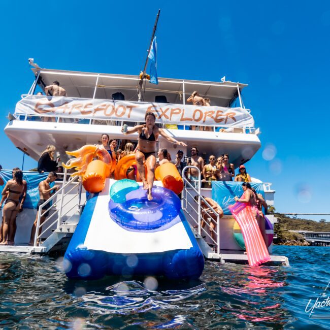 A group of people enjoying a sunny day on a boat named Barefoot Explorer. A woman slides down an inflatable water slide into the sea, while others relax on board. The logo "Yacht Social Club" is visible at the bottom right, highlighting Luxury Yacht Rentals in Sydney for unforgettable boat parties.