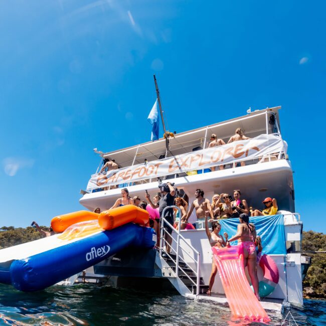 A group of people enjoying a sunny day on a two-story boat named "Barefoot Explore." Some are on the top deck, while others are gathered near a large slide that extends into the water. Inflatable floats are in use as the boat is anchored near a wooded shoreline, hosted by Boat Parties Sydney The Yacht Social Club.