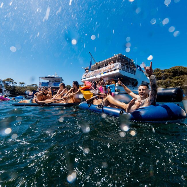 A group of people relax on inflatable floats and socialize in the water near a luxury yacht, enjoying a sunny day. The ocean is clear, and there are other boats and people in the background. Some individuals are splashing water, creating droplets in the air. Experience this with Yacht Social Club Sydney Boat Hire.