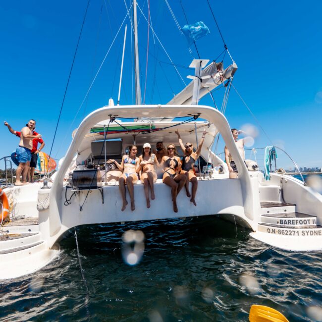 A group of people in swimwear sit on the edge of a white catamaran named "Barefoot," anchored under a clear blue sky. Some are waving and smiling at the camera, with scenic landscape and other boats visible in the background. Experience this moment with Luxury Yacht Rentals Sydney from The Yacht Social Club.