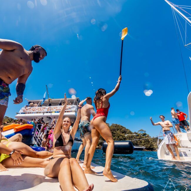 A group of people enjoying a sunny day on the water next to a luxury yacht. Some are sitting on a floating dock, while others are standing; one person is holding a paddle aloft. Everyone appears to be soaking up the sun and having fun in their swimwear, courtesy of Luxury Yacht Rentals Sydney.