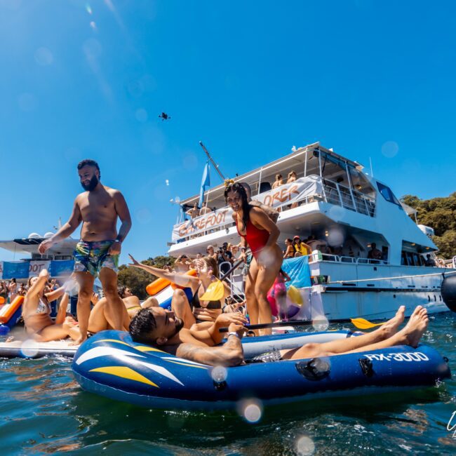 A group of people enjoy a sunny day on the water, some on inflatables and others standing, near a large white yacht with "The Yacht Social Club" signage. The vibrant scene includes colorful swimwear and a background of clear blue sky and greenery, epitomizing Boat Parties Sydney by The Yacht Social Club.