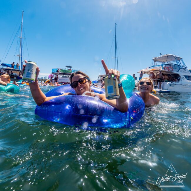 Two people are floating on an inflatable blue raft in the ocean, raising canned drinks in a celebratory manner. Boats and other people are visible in the background, suggesting a lively Yacht Social Club Sydney Boat Hire event. The sun is shining brightly, creating a festive, summery atmosphere.