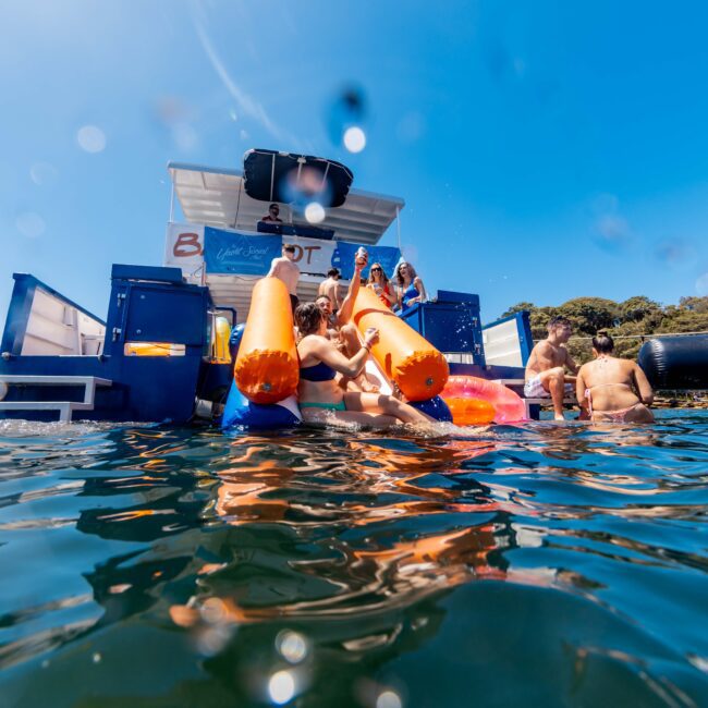 A fun scene on a sunny day where a group of people is gathered on a dock at the water's edge. They are enjoying various water toys and inflatables, with some people partially submerged in the water. A yacht is anchored nearby, showcasing "The Yacht Social Club Sydney Boat Hire.