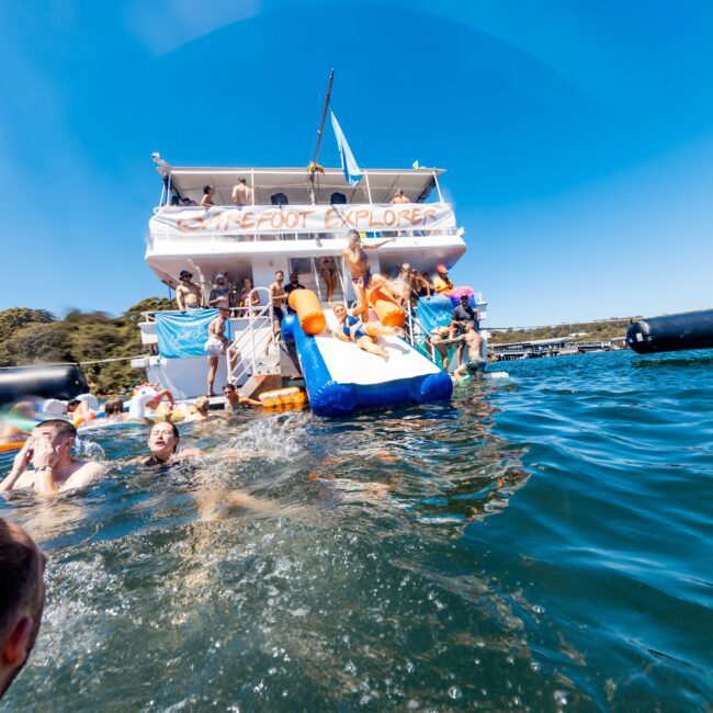 People are enjoying water activities around a two-story boat named "Barefoot Explorer" in a lake or ocean setting. The boat has a slide, and multiple floatation devices are visible. The sky is clear and the shore with trees is in the background, creating the perfect atmosphere for Boat Parties Sydney The Yacht Social Club.