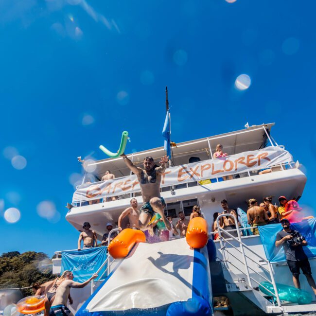 A group of people enjoy a sunny day on a double-deck yacht named "Yacht Explorer." Some are on the upper deck, while others go down an inflatable slide into the water. A person is mid-air jumping off the side, with a vibrant blue sky overhead, courtesy of The Yacht Social Club Sydney Boat Hire.