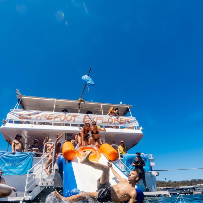 A group of people enjoy a sunny day on a boat labeled "Barefoot Explorer." One person is sliding down an inflatable slide into the water. The boat is on a clear blue lake with the bright sky above. 'Yacht Social' can be seen in the bottom right corner, reminiscent of Luxury Yacht Rentals Sydney.