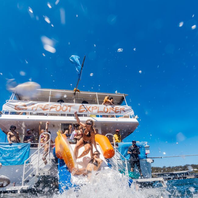 A group of people enjoy a sunny day on a two-level boat labeled "Barefoot Explorer," part of The Yacht Social Club Sydney Boat Hire. One person slides down an orange waterslide into the water, creating a large splash. Others cheer from the boat's upper deck, set against blue skies and a waterway backdrop.