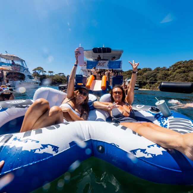 Two women relax on an inflatable float in the water, surrounded by other party-goers on floats and boats. They are smiling, wearing sunglasses, and holding drinks, with one making a peace sign. The scene is festive and sunny with clear blue skies and lush greenery—an ideal day at The Yacht Social Club Event Boat Charters.