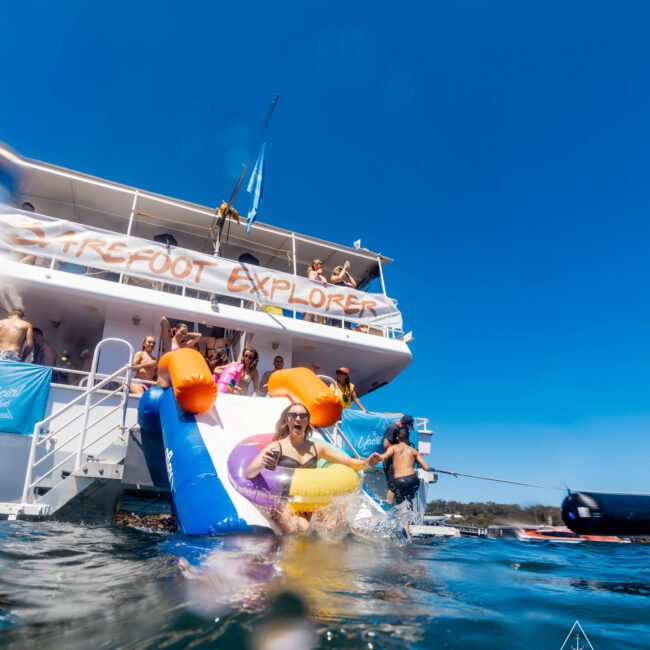 A person in a pink cap and bathing suit slides down an inflatable slide off a boat named "FreeSpirit Explorer" into the water. Other people stand on the boat, enjoying the sunny weather. The scene reflects bright blue hues, embodying quintessential Boat Parties Sydney by The Yacht Social Club.