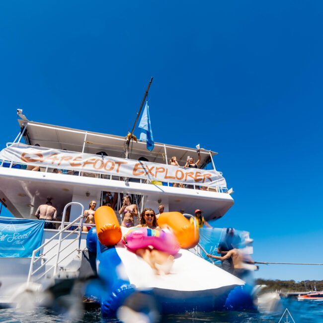 A person slides down an inflatable water slide into the ocean from a yacht named "Barefoot Explorer" with people on the upper deck watching. The yacht has a blue and white banner that reads "The Yacht Social Club," renowned for Boat Parties Sydney. The sky is clear and bright blue.