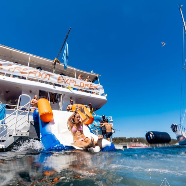 A group of people enjoys a sunny day on a yacht in Sydney Harbour. A woman in a pink bikini slides down an inflatable slide from the yacht into the water, with others watching and waiting their turn. The luxury vessel has "FRONT EXPLORER" written on its side, embodying the spirit of The Yacht Social Club.