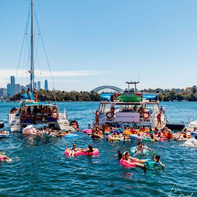 Two boats are anchored in a scenic waterfront area with a bridge and city skyline in the background. People float on colorful inflatables, enjoying a sunny day. The logo "Yacht Social" is visible in the bottom right corner of the image, showcasing Luxury Yacht Rentals Sydney from The Yacht Social Club.