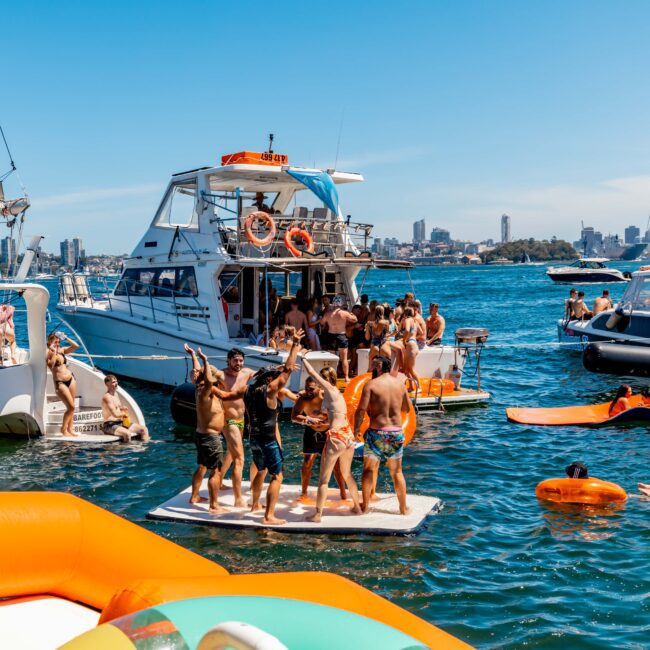 People enjoying a lively Yacht Social Club event on the water. Several boats are anchored close together, with partygoers dancing, swimming, and lounging on various floaties. The clear blue sky and distant city skyline form the backdrop of this Sydney Harbour Boat Hire experience.