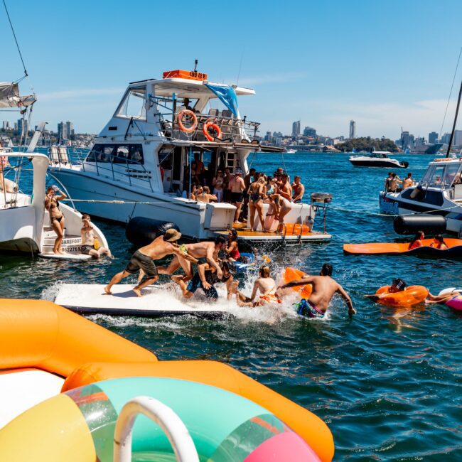 A lively scene of people enjoying a sunny day on a lake with several boats and inflatables from Sydney Harbour Boat Hire The Yacht Social Club. Some are playing and splashing in the water, while others relax on deck. The background shows a city skyline. The atmosphere is vibrant and festive.