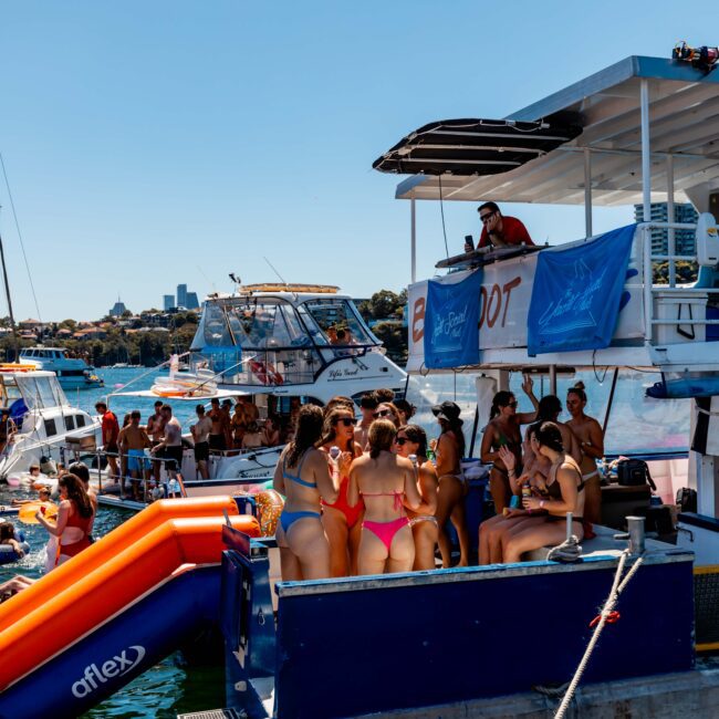 A lively scene of people enjoying a sunny day on the water. Several boats are anchored close together with people socializing, swimming, and lounging. Inflatable floats and a slide are visible. The atmosphere is festive, much like The Yacht Social Club's boat parties in Sydney with clear blue skies and calm water.