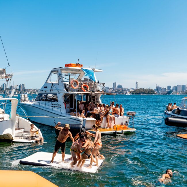 A lively scene of people enjoying a sunny day on a boat in open water, typical of The Yacht Social Club events. Individuals jump into the water while others socialize on the deck. Other boats and the stunning city skyline are visible in the background.