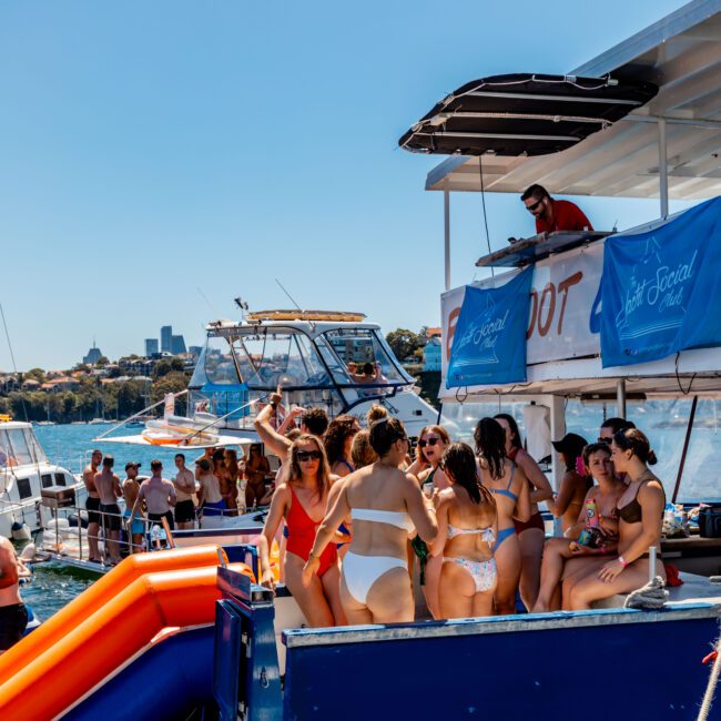 A group of people in swimwear stands on a yacht enjoying a party hosted by The Yacht Social Club, with several boats and a city skyline in the background. A bright inflatable slide extends from the yacht into the water. The scene is vibrant and sunny, depicting a lively summer atmosphere on Sydney Harbour.