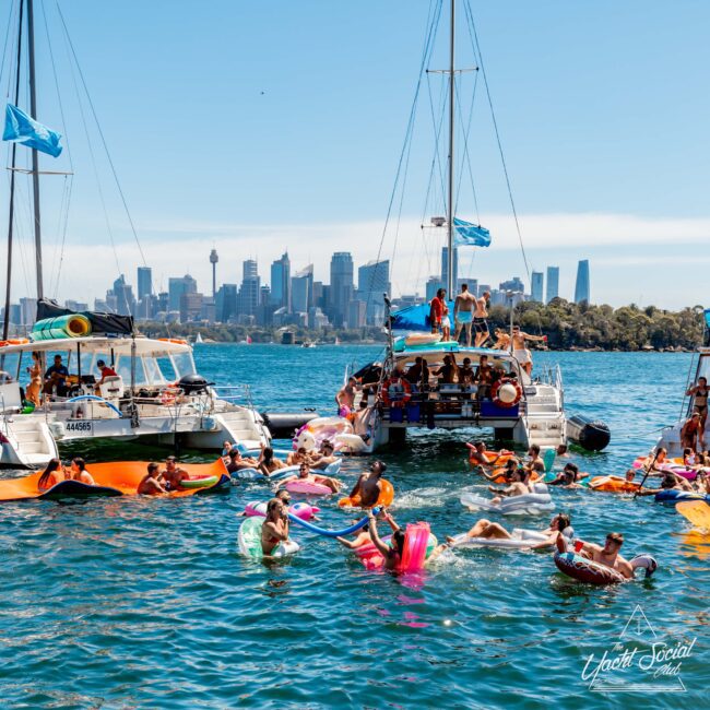 People enjoying a sunny day on the water, swimming and floating on various inflatable devices between anchored yachts, as part of The Yacht Social Club event. High-rise buildings of a city skyline visible in the background. Bright blue skies and vibrant water create a festive atmosphere.