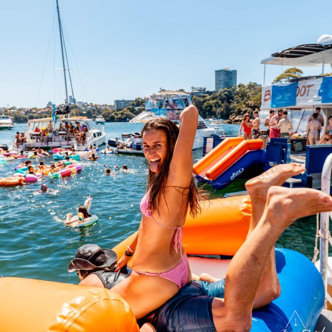 A woman in a pink bikini sits on an inflatable structure on a boat, smiling playfully with her arm raised. A man in a hat is behind her. Numerous people are enjoying the water and inflatables around other boats in the background. It’s a lively, sunny day at The Yacht Social Club Sydney Boat Hire.