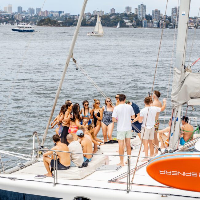 A group of people enjoying a day on "Barefoot," with a city skyline and other sailboats in the background. The weather is clear, and everyone appears relaxed and engaged in conversation. Consider joining Sydney Harbour Boat Hire for more such experiences with The Yacht Social Club.