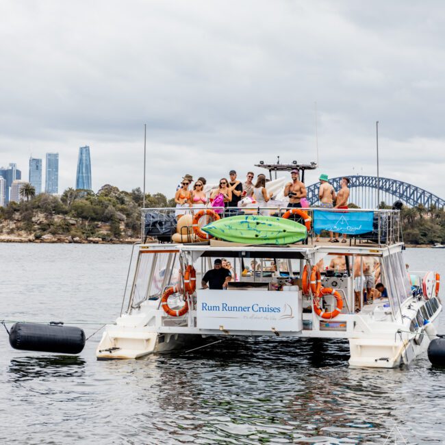 A group of people enjoy a boat cruise on a catamaran named "Rum Runner Cruises," surrounded by scenic water with a cityscape and the Sydney Harbour Bridge in the background. Paddleboards are secured on the roof, with life buoys attached to the sides. Experience this with Sydney Harbour Boat Hire at The Yacht Social Club.