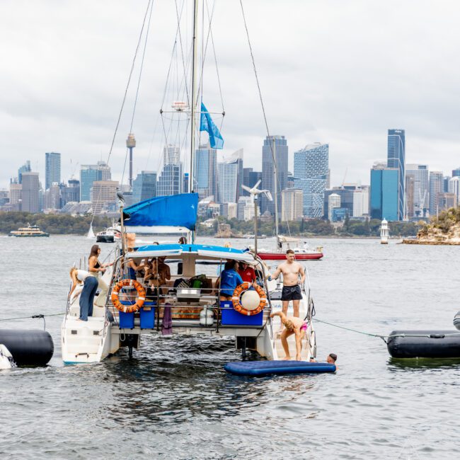 A group of people enjoy a day on a sailboat anchored in Sydney Harbour with the city skyline in the background. Some individuals are on the boat, while others are in the water and on a floating platform, partaking in The Yacht Social Club event. The sky is overcast.