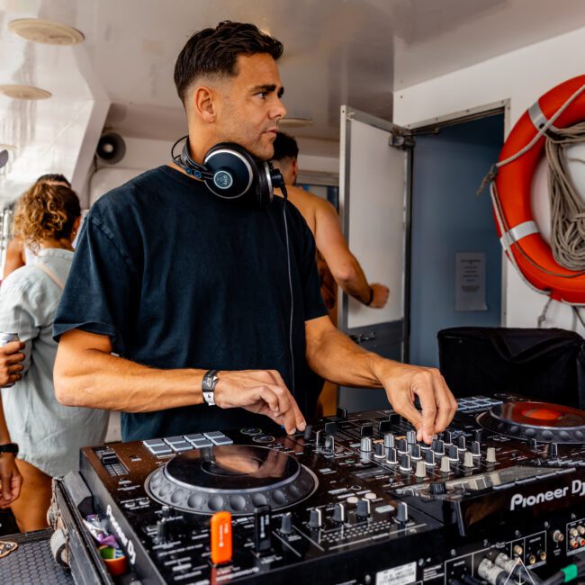 A DJ in a black shirt and headphones stands behind a Pioneer DJ controller, adjusting the equipment with his hands. The setting appears to be an indoor area possibly on a boat, evidenced by the life preserver on the wall. People in casual attire are in the background, enjoying The Yacht Social Club Sydney Boat Hire.