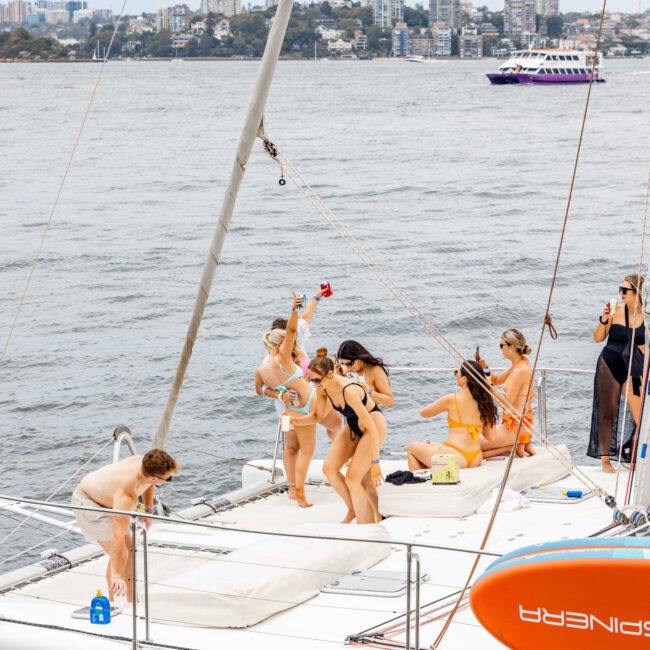 A group of people enjoying a day on a yacht. Some are standing, with one person raising a drink, while others are sitting or lying down on the deck. The yacht is sailing on water with a cityscape in the background during The Yacht Social Club Event Boat Charters. A purple and white boat is also visible in the distance.