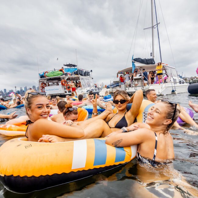 A group of people relax on colorful inflatable floats in the water near a boat. The overcast sky and several other boats in the background suggest a festive gathering or party, typical of The Yacht Social Club Event Boat Charters. People are smiling and enjoying the event.