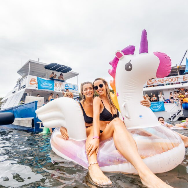 Two people in swimsuits sit on an inflatable unicorn float in the water, smiling and posing for the camera. In the background, there are boats with people, including one with the sign "BAREFOOT BLUE." The vibrant scene appears to be a lively summer event hosted by Boat Parties Sydney The Yacht Social Club.