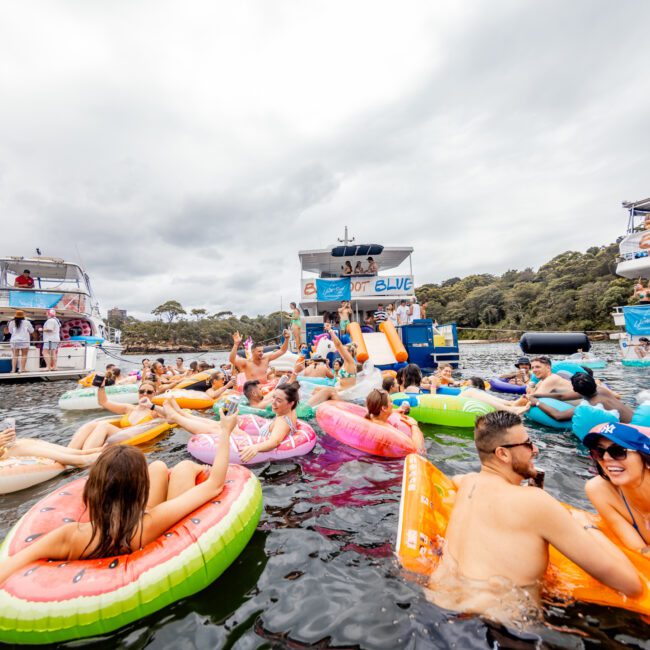 A lively scene of people enjoying a party on a lake. Many are floating on colorful inflatable rafts while others swim around. Two boats with banners and music from The Yacht Social Club are in the background, against an overcast sky. Everyone seems to be having fun and socializing.