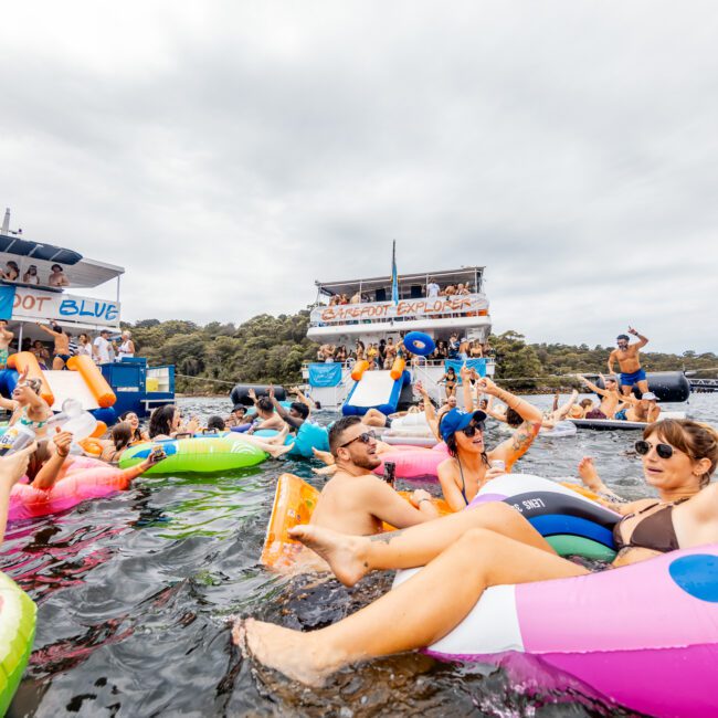 A lively scene on the water shows people enjoying a party. Individuals float on colorful inflatables, mingling and laughing. In the background are two anchored boats with people onboard, courtesy of Sydney Harbour Boat Hire The Yacht Social Club. The sky is cloudy, and the atmosphere is festive and joyful.