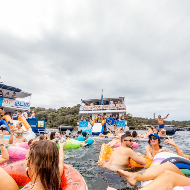 A lively scene of a boat party by The Yacht Social Club. People float on inflatable rafts, socializing and enjoying the day. The boat in the background is decorated with banners and has a slide. The atmosphere is festive with overcast skies and lush greenery behind.