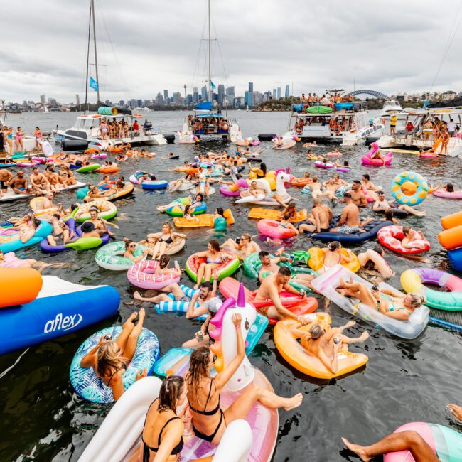 A lively scene on a body of water with numerous people floating on colorful inflatable rafts and pool toys. Boats are anchored in the background under a cloudy sky, with a city skyline visible in the distance. The Yacht Social Club Event Boat Charters hosts a festive atmosphere filled with joy and entertainment.