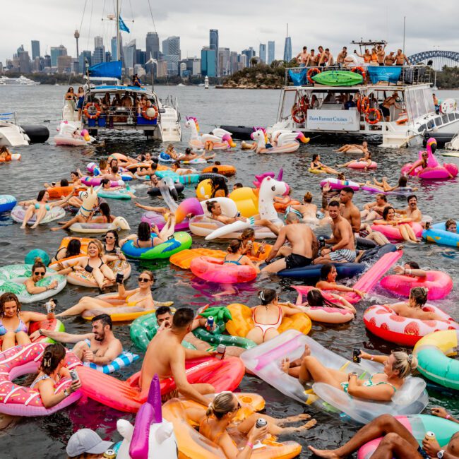 A large group of people on colorful inflatable floats, including unicorns and donuts, are gathered and relaxing in the water near several anchored boats. The city skyline is visible in the background under a cloudy sky. The Yacht Social Club Sydney Boat Hire adds a vibrant and festive touch to the scene.