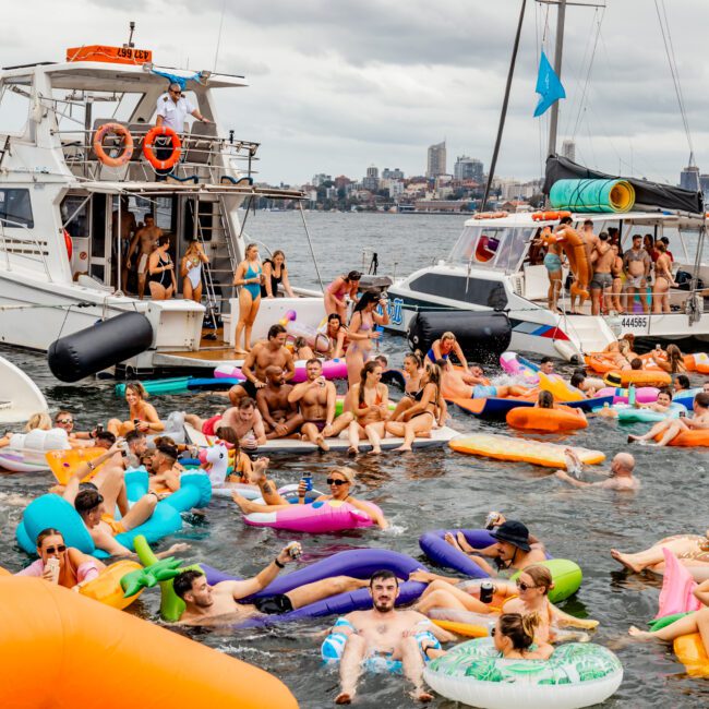 A lively scene of people enjoying a boat party on the water, with many individuals floating on colorful inflatable tubes and rafts. Several boats from The Yacht Social Club Sydney Boat Hire are anchored nearby, and people on the boats are socializing under a cloudy sky.