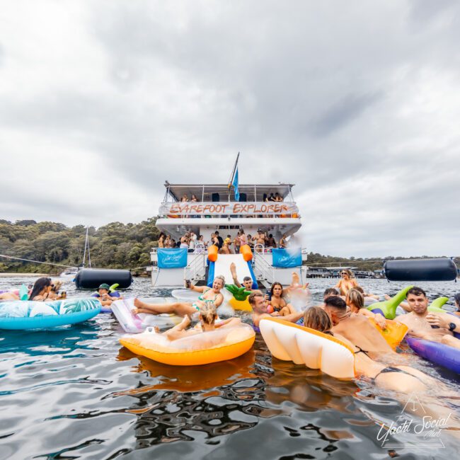 A group of people float on colorful inflatables in a body of water near a large boat named "Barefoot Explorer." Others are on the boat, with "The Yacht Social Club Event Boat Charters" written on its side. The sky is overcast, and the scene is lively with individuals enjoying the water.