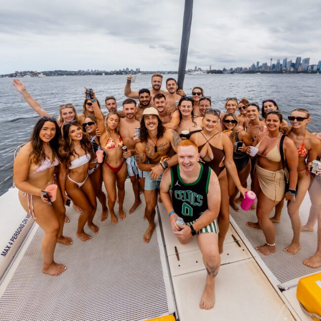 A group of people in swimsuits is gathered on the deck of a boat from The Yacht Social Club, smiling and posing for a photo. They are on the water with a city skyline visible in the background under a cloudy sky. Some are holding drinks, creating a joyful and celebratory atmosphere.